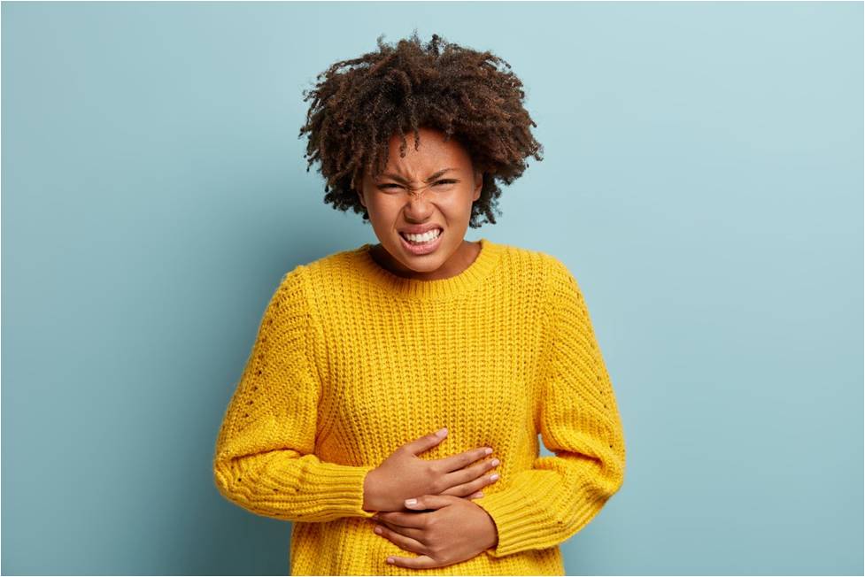 A young black woman in a yellow sweater holding her belly with both hands, showing discomfort due to period pain.