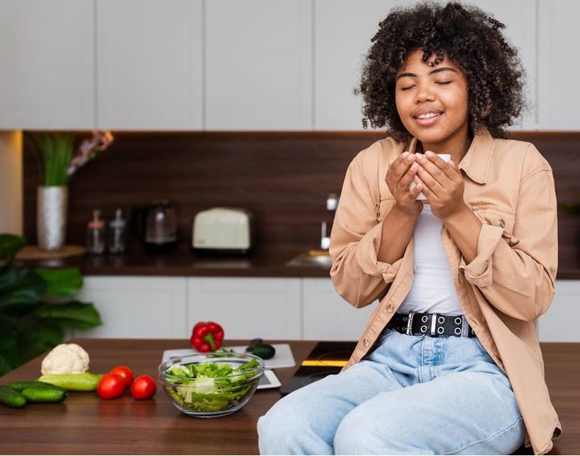 A young woman with curly hair sitting on a kitchen counter, holding a cup and enjoying the aroma of her drink, surrounded by fresh vegetables